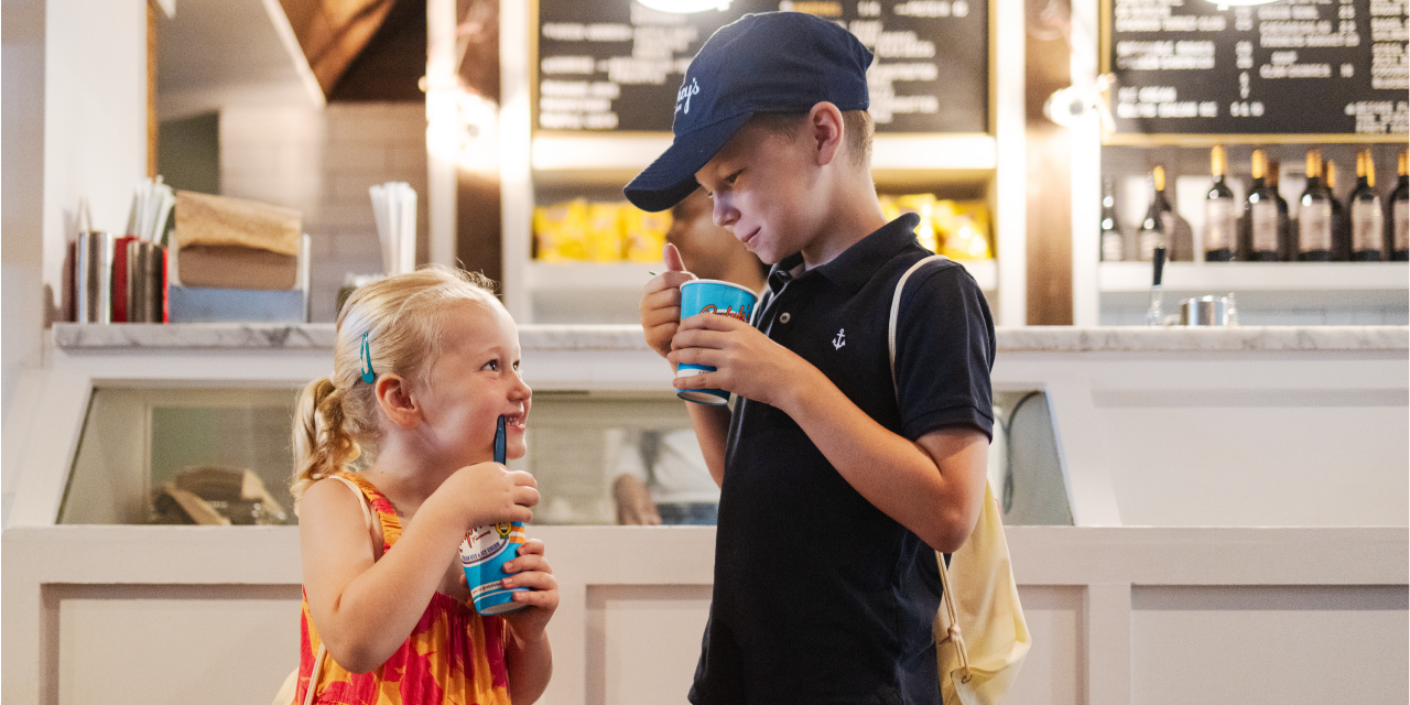 Two children stand in front of a counter holding cups of ice cream and smiling