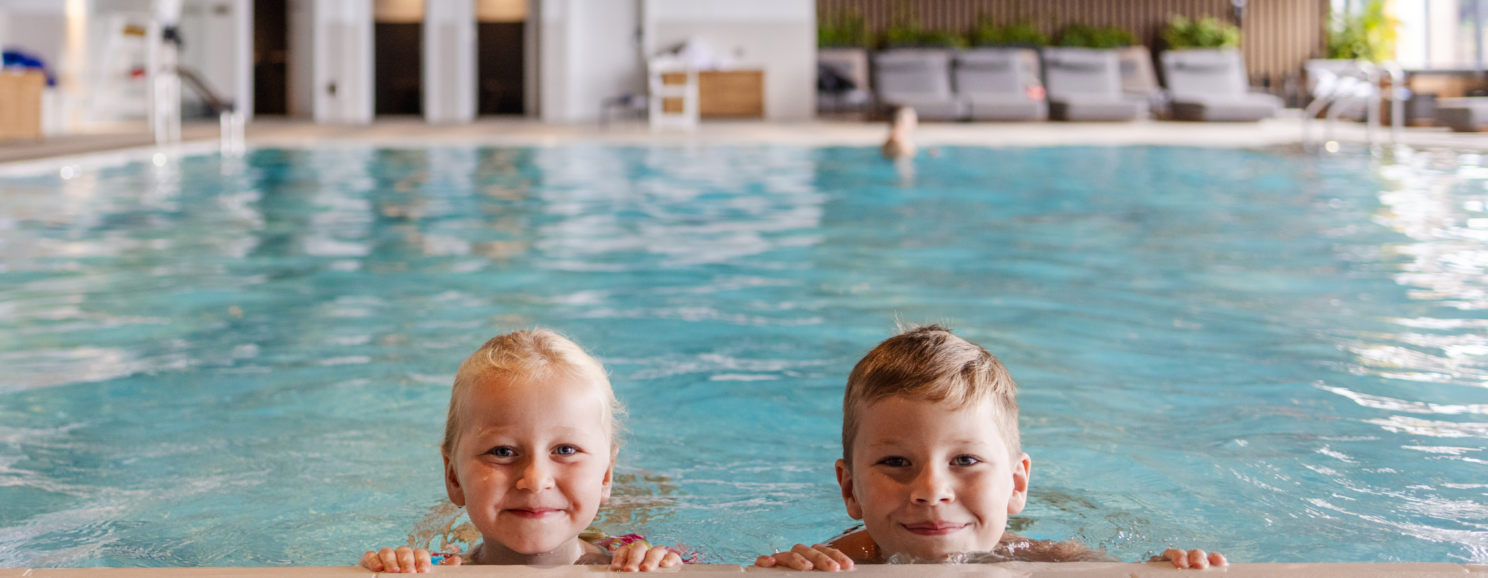 Two children smile at the edge of an indoor pool