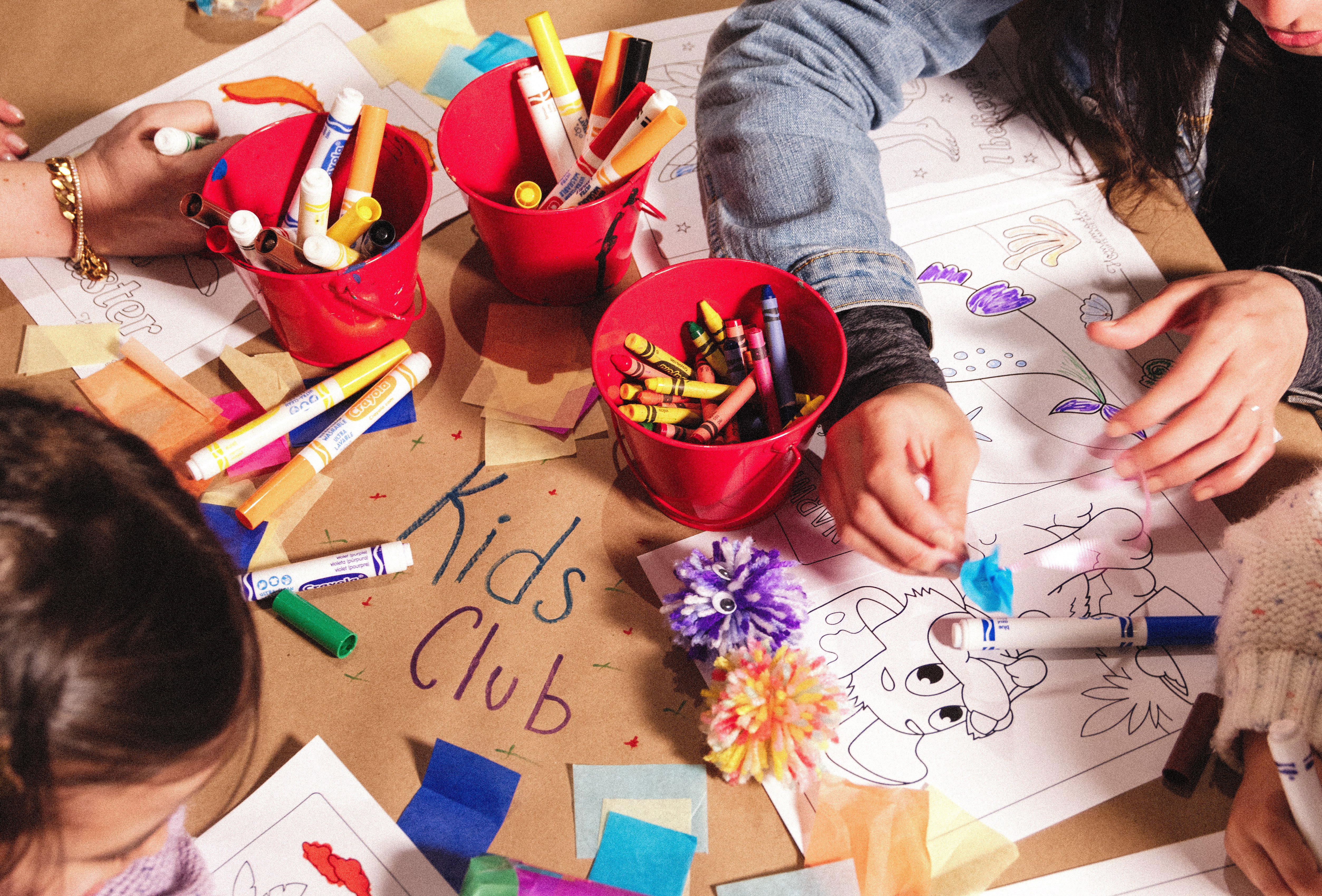 Children coloring at a table with red cups of markers and crayons, surrounded by paper and crafts. Brown paper reads "Kids Club."