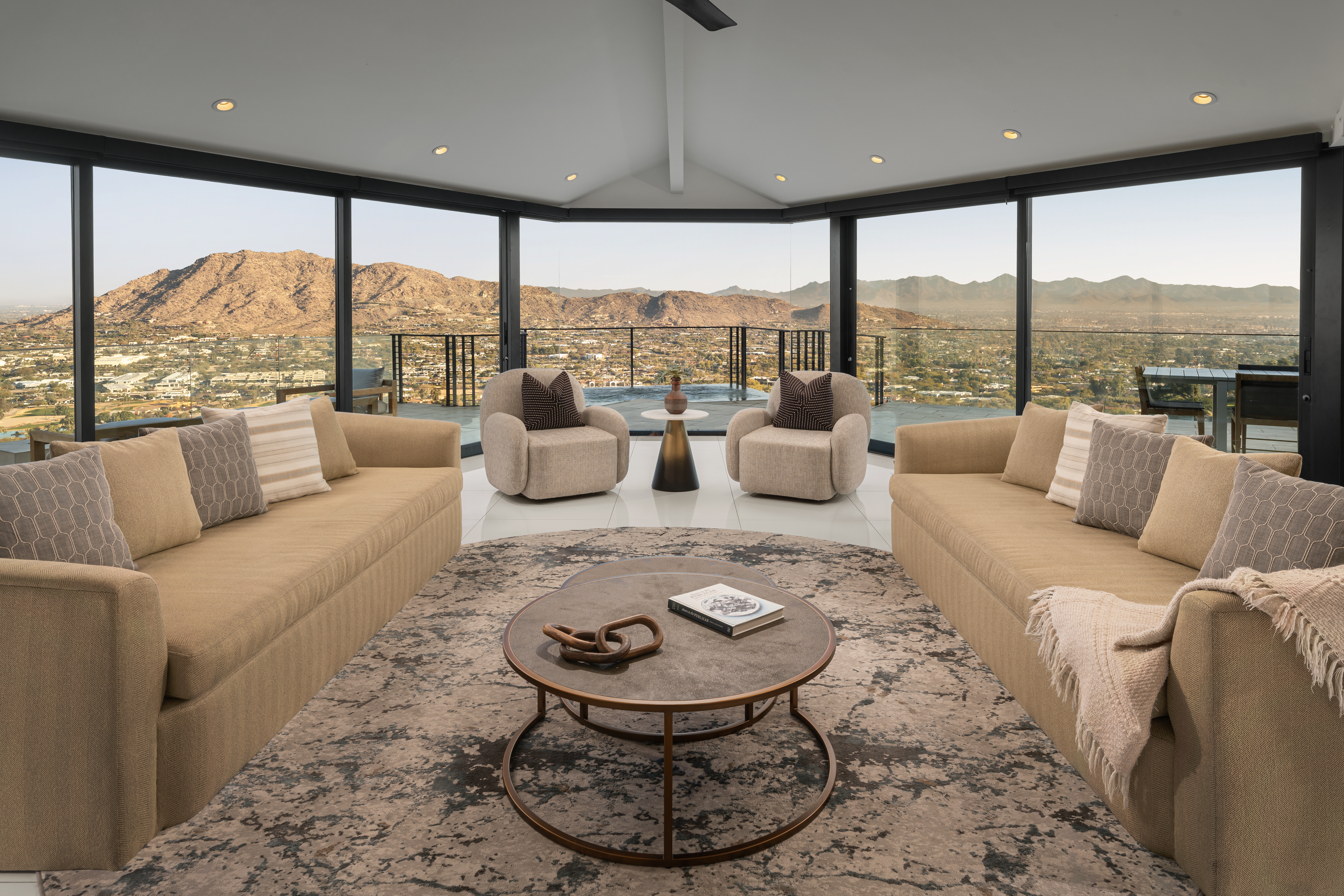 Elegant living room with large windows, mountain view, two beige sofas, two armchairs, and a round coffee table on a patterned rug.