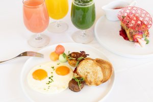 Breakfast with sunny-side-up eggs, toast, and fruits. Three colorful juice glasses. Dessert with red netting decoration on a separate plate.