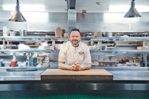 Chef in a white uniform leans on a wooden counter in a bustling, stainless steel kitchen.