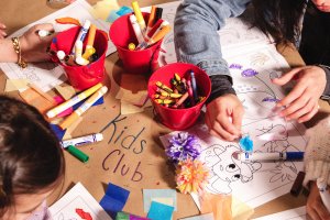 Children coloring at a table with red cups of markers and crayons, surrounded by paper and crafts. Brown paper reads "Kids Club."