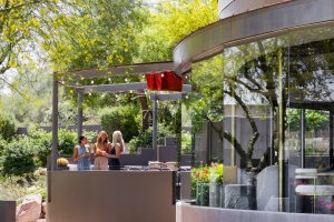 Modern outdoor café with people seated at tables, surrounded by lush greenery and string lights. Reflective glass facade curves on the right.