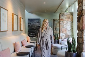 Woman in a bathrobe walks through a spa lounge with soft lighting, stone walls, and secluded seating.