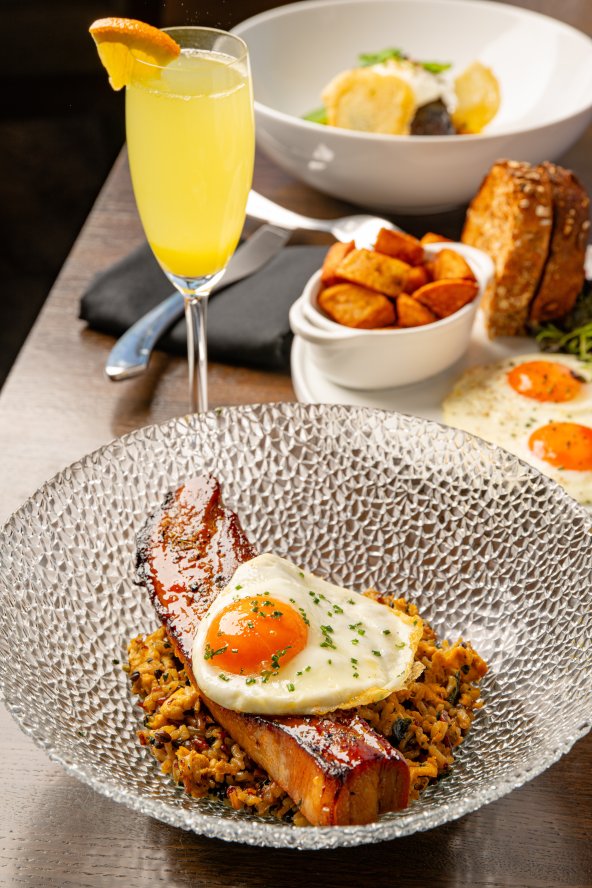 Breakfast dish with a fried egg on bacon and hash, accompanied by a mimosa, bread, and fruit bowl on a wooden table.