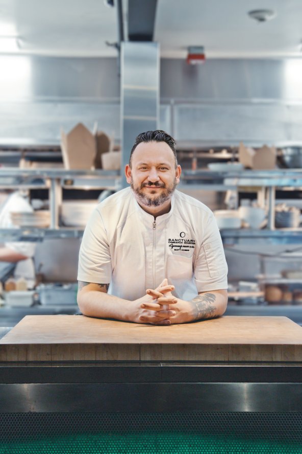 Chef in a white uniform leans on a wooden counter in a bustling, stainless steel kitchen.