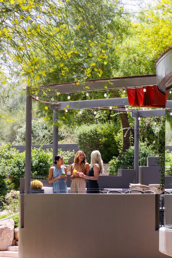 Modern outdoor café with people seated at tables, surrounded by lush greenery and string lights. Reflective glass facade curves on the right.