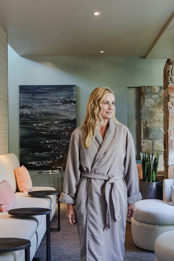 Woman in a bathrobe walks through a spa lounge with soft lighting, stone walls, and secluded seating.