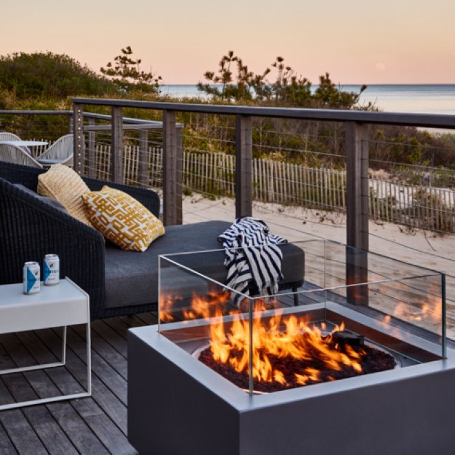 Deck with comfortable lounge chair and fire pit with view of beach at sunset.