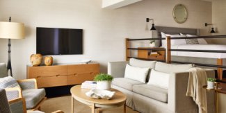 Cozy living room with beige sofa, small coffee table, wall-mounted TV, and modern decor. Bed on loft above, neutral tones throughout.