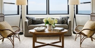 Cozy living room with large windows, two armchairs, a gray sofa, a round wooden table with white flowers, and two floor lamps. Ocean view in the background.