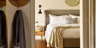 Cozy bedroom with a neatly made bed, gray headboard, pendant light, side table, and wall mirror; blankets and a cushion add warmth.