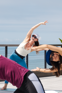 Women in colorful athletic clothes bend in a yoga pose in a guided class with a view of the ocean in the background