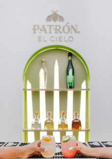 Two hands hold brightly colored cocktails against a shelf with Patron tequila